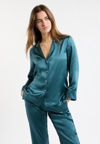 Conjunto de pijama de satén color verde azulado que incluye una blusa abotonada con cuello y mangas largas, combinada con pantalones de pierna ancha a juego. Textura suave y brillante.
