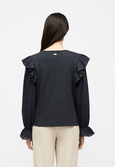 Femme aux longs cheveux porte un haut noir à manches longues avec des épaules volantées et des poignets ornés de dentelle, associé à un pantalon beige clair, vue de dos.