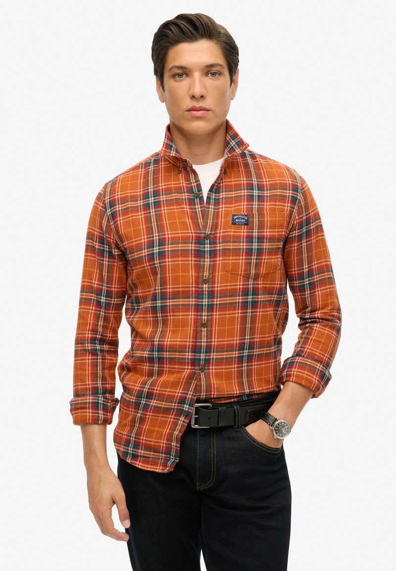 Camisa de cuadros en naranja, azul y verde, con cuello abotonado y bolsillo en el pecho. Combinada con jeans oscuros y un cinturón negro.