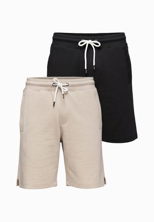 Homme debout portant un T-shirt blanc avec du texte, un short noir, des baskets blanches et un bracelet noir au poignet gauche.