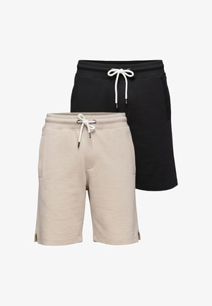 Mann steht, trägt ein weißes T-Shirt mit Text, schwarze Shorts, weiße Turnschuhe und ein schwarzes Armband am linken Handgelenk.