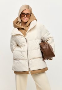 Kobieta o krótkich blond włosach, ubrana w beżową puchową kurtkę na brązowej bluzie z kapturem, beżowe spodnie, okrągłe, duże okulary przeciwsłoneczne, trzymająca brązową torebkę.