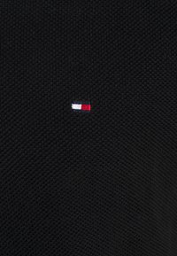 Kleines gesticktes rechteckiges Logo mit weißen, roten und marineblauen Abschnitten auf schwarzem strukturiertem Stoff.