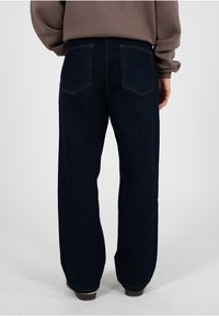 Person trägt dunkelblaue, gerade geschnittene Jeans und ein braunes Langarmshirt, steht auf einem schlichten hellen Hintergrund mit dem Rücken zum Betrachter.