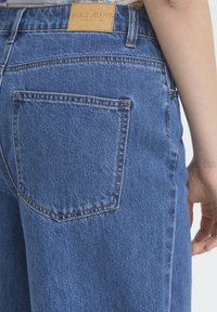 Jean en denim bleu avec une poche arrière rectangulaire et une étiquette en cuir marron portant l'inscription "PULZ JEANS". Caractérisé par des coutures visibles et une coupe décontractée.