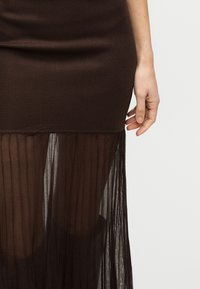 Το χέρι μιας γυναίκας που ακουμπάει δίπλα σε μια σκουρόχρωμη καφέ εφαρμοστή φούστα με διαφανή, πιετέ χαμηλή περιοχή, πάνω σε ένα απλό φόντο.