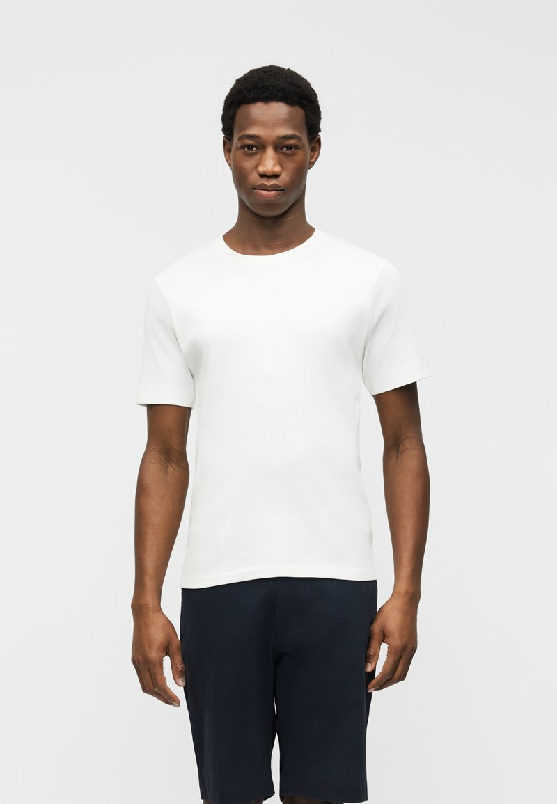 Uomo in piedi, volto frontale, indossa una maglietta bianca a maniche corte semplice e pantaloncini scuri, su sfondo grigio chiaro.