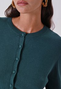 Dunkelgrüner Cardigan mit Rundhalsausschnitt, mit Knopfverschlüssen vorne und einem weichen, strukturierten Stoff.