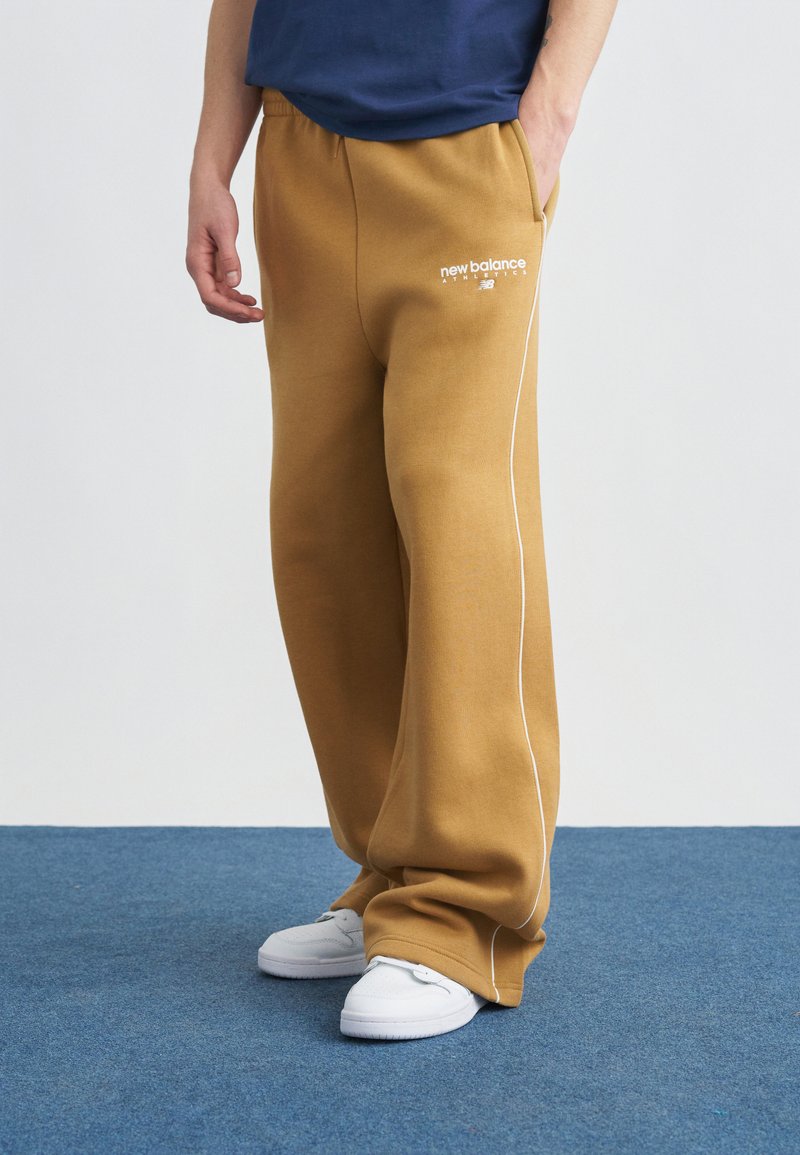 Άτομο που φοράει μουσταρδί παντελόνι φόρμας New Balance με λευκό ρέλι και λευκά αθλητικά παπούτσια, στέκεται σε μπλε χαλί.