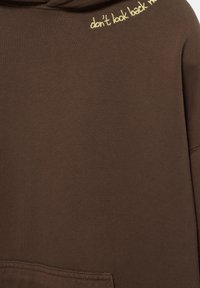 Hnedá mikina s jemnou textúrou, so žltým vyšitým nápisom "nedívaj sa späť" pozdĺž výstrihu a prednou vreckom.