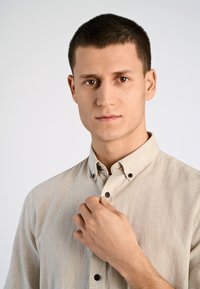 Camisa de botones color beige claro con una tela texturizada, que presenta botones oscuros. El cuello es clásico y el ajuste parece ser entallado.