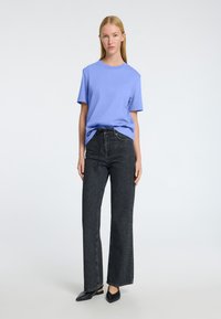 T-shirt crew en violet clair associé à un jean noir taille haute à jambes larges et des mules noires pointues, sur fond uni.