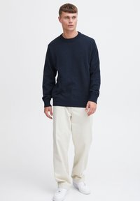 Maglione a maglia blu navy con finitura texture, colletto a girocollo e polsini a costine, abbinato a pantaloni di colore chiaro e sneakers bianche.