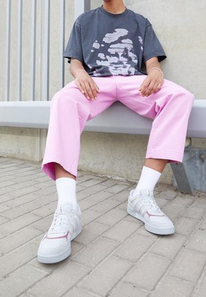 Persona seduta su una panchina grigia che indossa una t-shirt grafica grigio scuro, pantaloni larghi rosa, calzini bianchi e sneaker bianche con dettagli rosa.