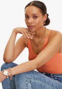 Roséguld kædedyse, matchende armbånd og ring; model iført en orange ribbet tanktop og denim jeans; glat hudtekstur.