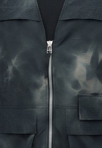 Μπροστινή κοντινή λήψη ενός σκούρου μπουφάν με κεντρικό ασημί φερμουάρ και δύο μεγάλες τσέπες με καπάκι στο κάτω μισό.