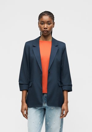 Fonott hajú nő sötétkék blézerben, narancssárga inggel és világoskék gyöngyös díszítésű farmerral, egyszínű háttér előtt állva.