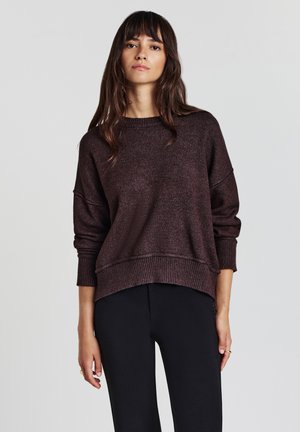 Felpa in maglia bronzo con una vestibilità rilassata, maniche larghe e orlo a costine. Abbinata a pantaloni neri aderenti. Texture morbida, design casual.