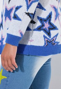 Gebreide trui met een blauw sterpatroon op een witte achtergrond, geribbelde boorden en zoom, gecombineerd met lichtblauwe jeans met een gele sterdetail.