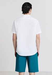 Camicia bianca a maniche corte con colletto alla francese, abbinata a pantaloni corti azzurri fino al ginocchio. Il tessuto appare liscio e leggero.