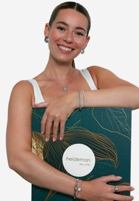 Boîte à bijoux avec un design vert foncé orné de feuilles dorées et d'un logo blanc. Le modèle la tient en montrant des boucles d'oreilles, un collier et des bracelets en argent.