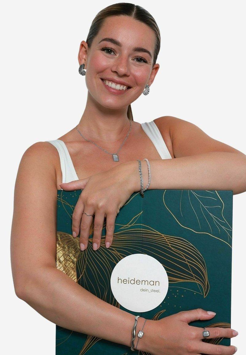 Boîte à bijoux avec un design vert foncé orné de feuilles dorées et d'un logo blanc. Le modèle la tient en montrant des boucles d'oreilles, un collier et des bracelets en argent.
