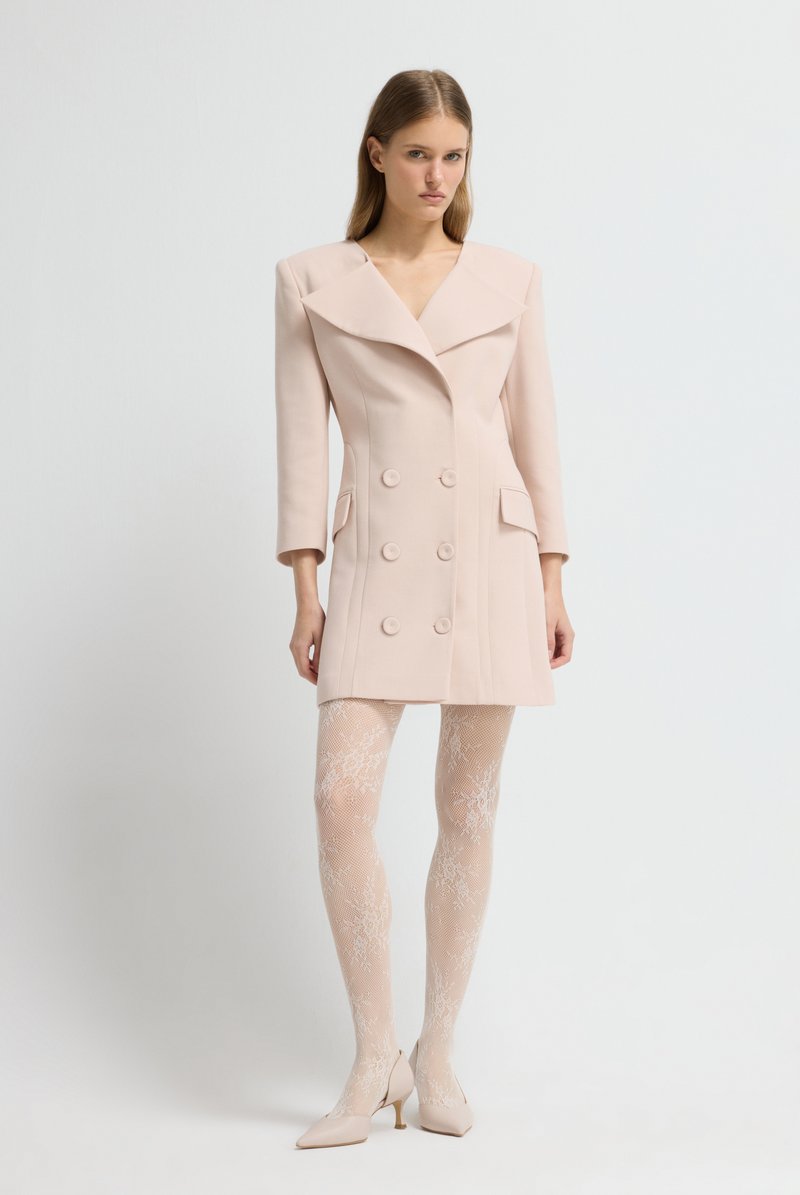 Cappotto doppio petto rosa chiaro con ampie rever e due tasche frontali, abbinato a calze color nude a fantasia e tacchi di colore chiaro.