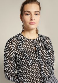 Schwarze transparente Bluse mit weißem gewelltem Muster, runder Ausschnitt, Rüschen vorne und langen Ärmeln.