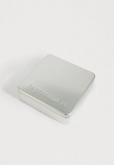 Mažas sidabrinis stačiakampis metalinis dėklas su apvaliais kampais, išspausdintu užrašu "le gramme" ir mažu kvadratu ant dangtelio, ant šviesios fono.