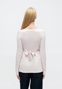 Hosszú ujjú, rózsaszín bordázott felső, testhezálló szabással, derékrészen szatén masnival díszítve, sötét farmernadrággal párosítva.