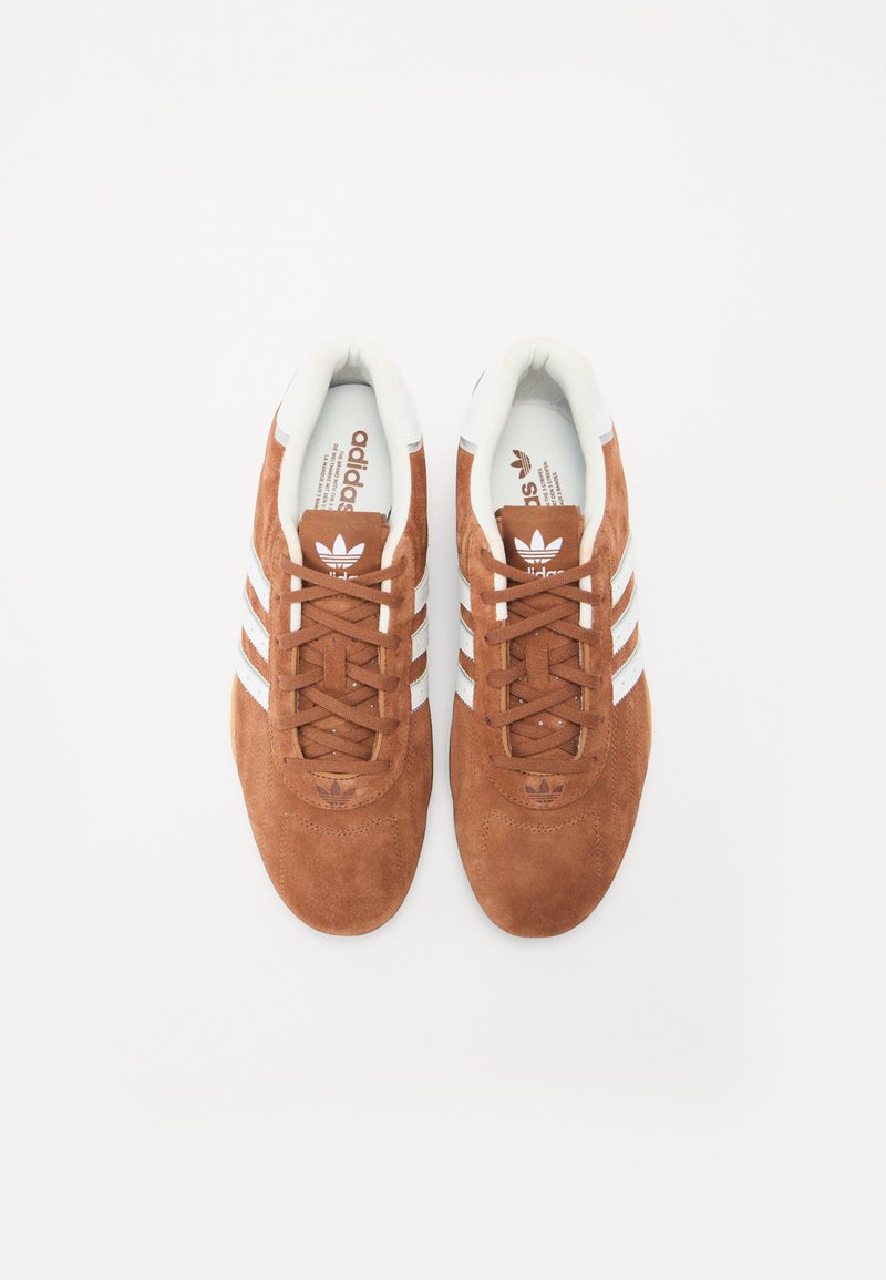 Braune Wildledersneaker mit weißen Drei-Streifen-Akzenten, runder Zehenpartie, flachen Schnürsenkeln und einer gepolsterten Zunge für zusätzlichen Komfort.