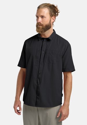 Baarddragende man met naar achter gebonden haar, gekleed in een zwart overhemd met korte mouwen en lichte broek, licht naar links gewend.