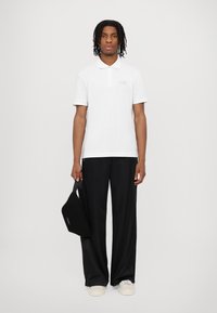White polo shirt with short sleeves and a subtle logo, paired with loose black trousers and a small black bag, worn with beige shoes.