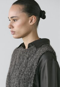 Gray knitted vest made of textured fabric, layered over a black collared shirt. Side profile view, highlighting the garment's materials and shape.