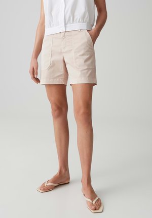 Person wearing light beige shorts with large front pockets, a white sleeveless buttoned top, and cream flip-flops, standing with one hand in pocket.