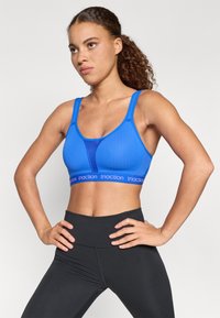 Modrý športový top s sieťovým dizajnom, širokými ramienkami a elastickým pásom so značkou "triaction". V páre s čiernymi legínami s vysokým pásom.