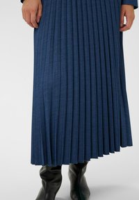 Kéz a világoskék háttéren egy sötétkék, pliszírozott midi szoknya mellett, fekete, hegyes orrú bőr csizmákkal párosítva.