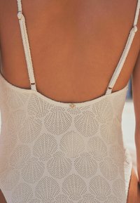 Maillot de bain en crochet blanc avec des motifs en coquille, bretelles réglables et un petit accent doré dans le dos. Texture douce et coupe ajustée.