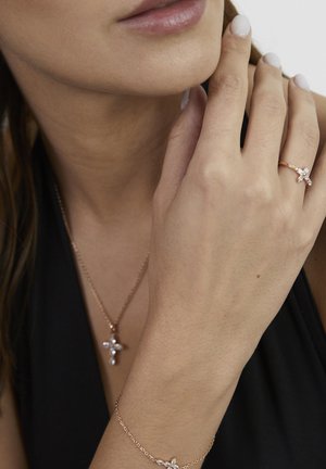 Close-up of a woman wearing a cross-shaped ring, matching necklace, and bracelet with clear gemstones, hand touching her face.