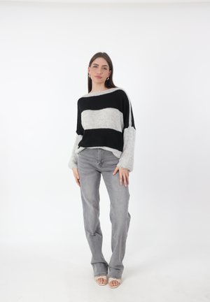 Maglione a righe nero e grigio con una vestibilità ampia e maniche larghe, abbinato a jeans grigio chiaro e sandali di colore chiaro, su uno sfondo semplice.