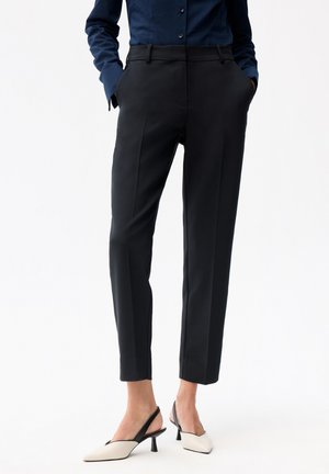 Personne portant un pantalon noir sur mesure avec des poches et des chaussures à talons kitten noires et blanches à bride arrière, debout devant un fond blanc.