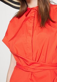 Kvinna i en ljusorange klänning med krage, knytning i midjan och lösa korta ärmar, delvis sedd med brunt hår och en halmhatt.