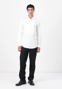 Witte lange mouwen blouse met een geplooide voorkant, gecombineerd met zwarte broek en glanzende zwarte schoenen, geplaatst tegen een effen achtergrond.