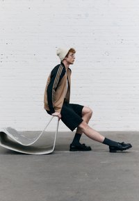 Urheilutakki ruskean ja mustan sävyissä, jossa on kolme raitaa, shortsit, mustat loaferit ja beige pipo, istumassa kaarevalla metallisella rakenteella.