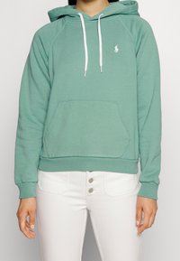 Sudadera con capucha verde en material suave, con un bolsillo frontal, cordones blancos y un pequeño logo en el lado izquierdo del pecho.