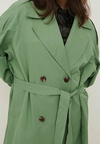 Kobieta w jasnozielonym płaszczu z paskiem noszonym na czarną skórzaną koszulę zapinaną na guziki, pokazana od szyi do połowy uda.