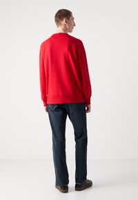 Rød sweatshirt med rund halsudskæring og lange ærmer, parret med mørk denim jeans og sorte sko. Modellen står i profil.