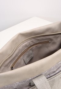 Interno di una borsa color beige che mostra una tasca con zip e fodera in tessuto con bordi rinforzati, poggiata su una superficie bianca.
