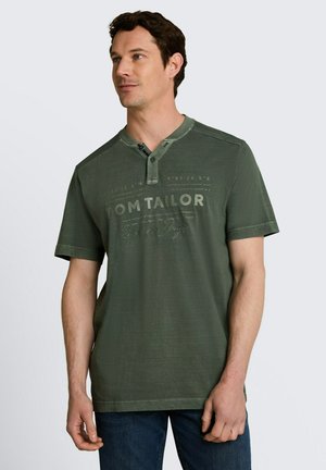 Mężczyzna w zielonej koszulce henley z krótkim rękawem z napisem „Tom Tailor” na piersi, stojący na jednolitym tle.