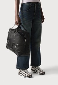 Borsa da viaggio nera realizzata in materiale sintetico con manici robusti, caratterizzata da una finitura lucida e dettagli con logo. Indossata con jeans a gamba larga in denim blu e scarpe.